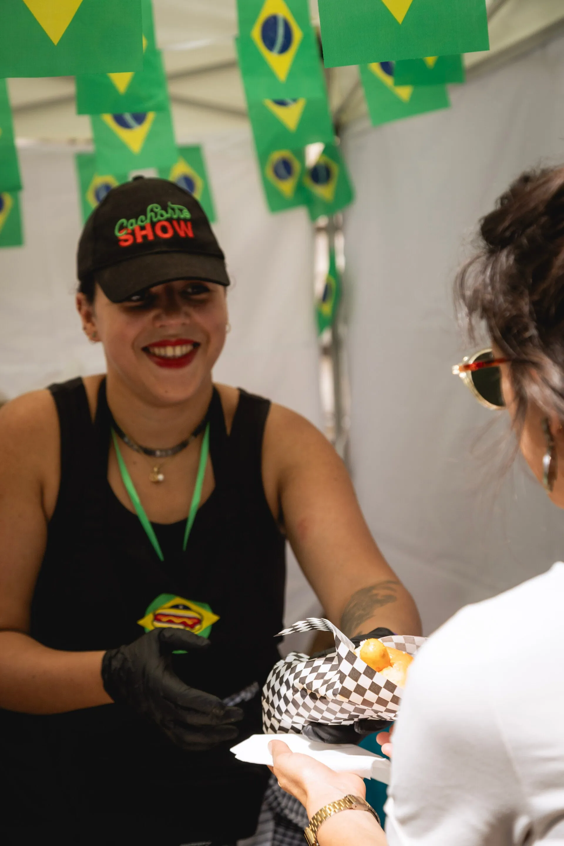 Monah, fondatrice de Cachorro Show, souriante derrière son stand décoré de drapeaux brésiliens